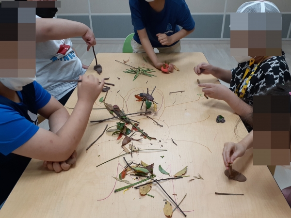 학생 5명이 책상에 둘러 앉아 점토에 나뭇가지, 풀잎 등을 사용하여 곤충을 만들고 있다.